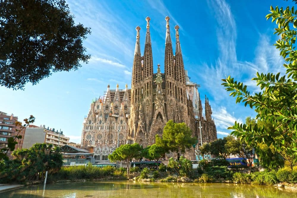 Ornate basilica with tall spindle-shaped spires rising above a park with trees and a pond