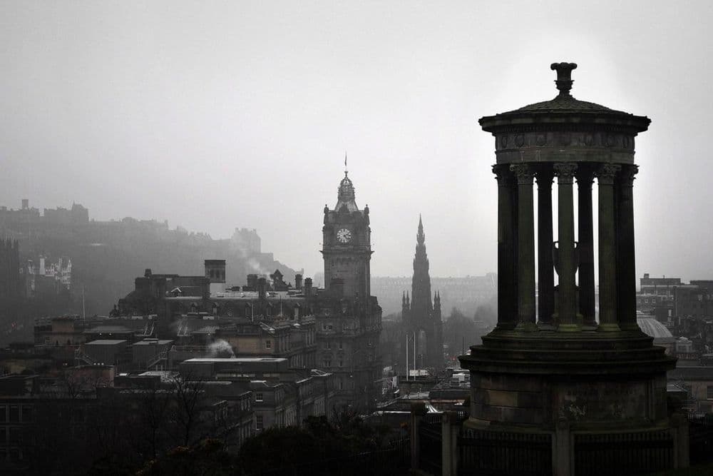 Round columned monument on a hill above a hazy city with a clock tower behind