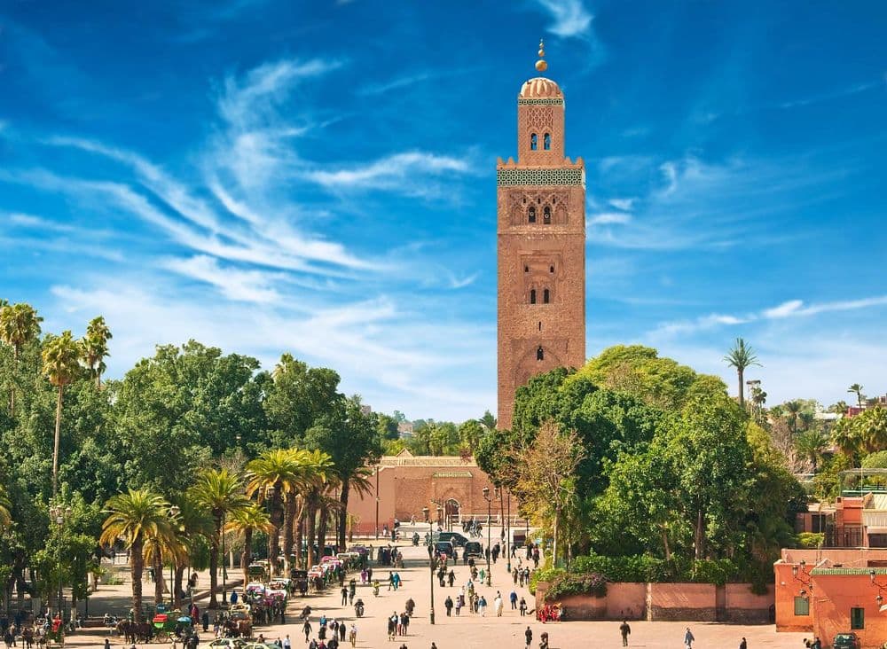 Minareto color arenaria dietro palme sopra una piazza affollata di persone